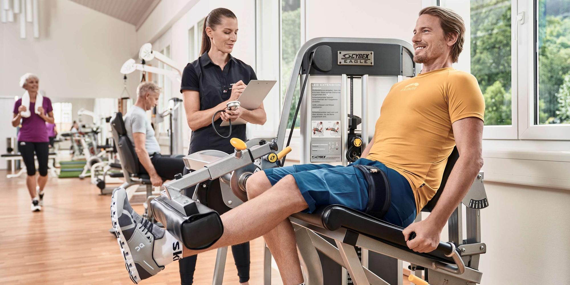 A man in a yellow t-shirt and blue shorts seated in Cybex Eagle Leg Press Machine under the guidance of a professional lady in black t-shirt. A woman in a black t-shirt with and a man in a yellow t-shirt and blue shorts seated in Cybex Eagle Leg Press Machine.