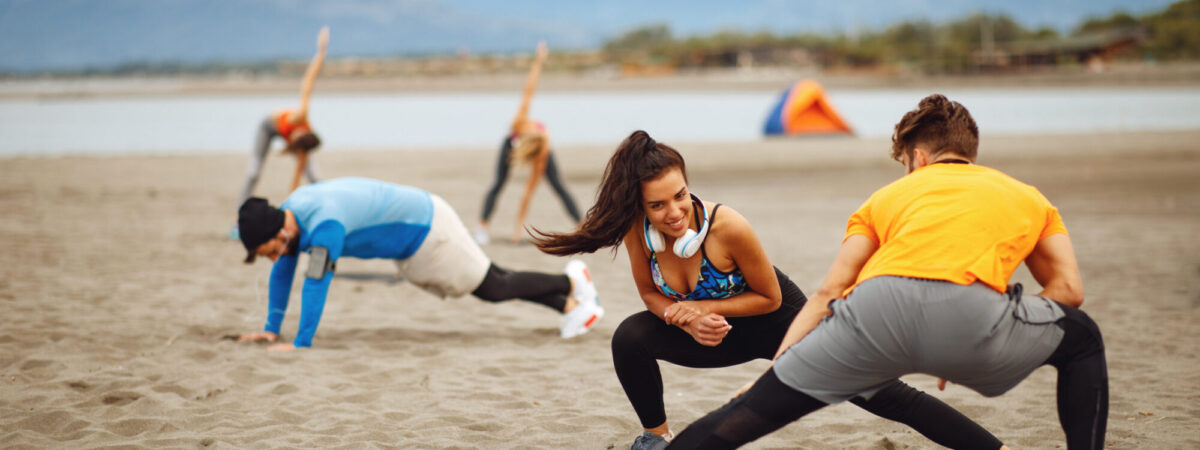 People in athletic wear doing lunges and stretches on a sandy beach, with mountains in the background and a colorful tent nearby.