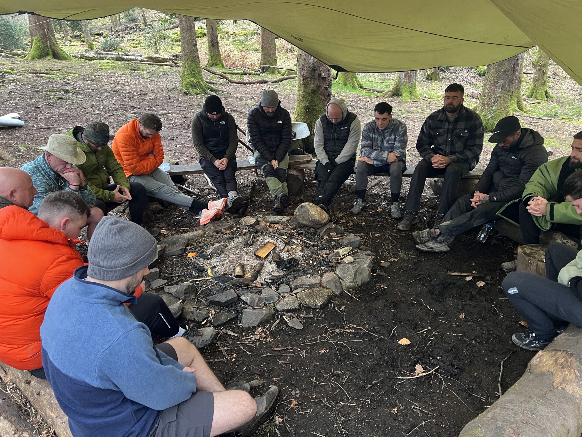 An image of individuals participating in an outdoor activity, likely a hiking or wilderness training session. Group of people sitting in a circle around a small campfire under a tarp in a forest area.