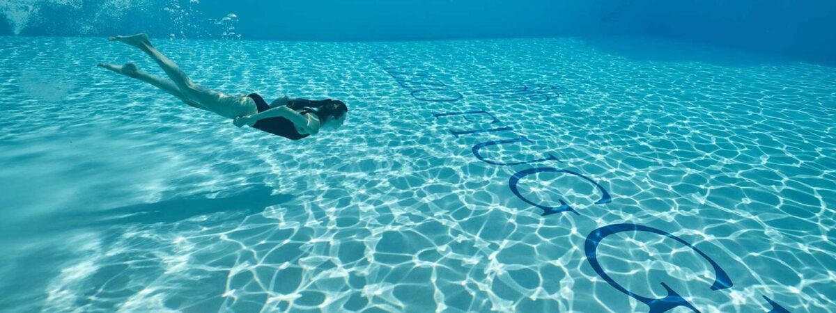Person swimming underwater in a clear blue pool, side view, with sunlight patterns on the pool floor, bubbles ahead.