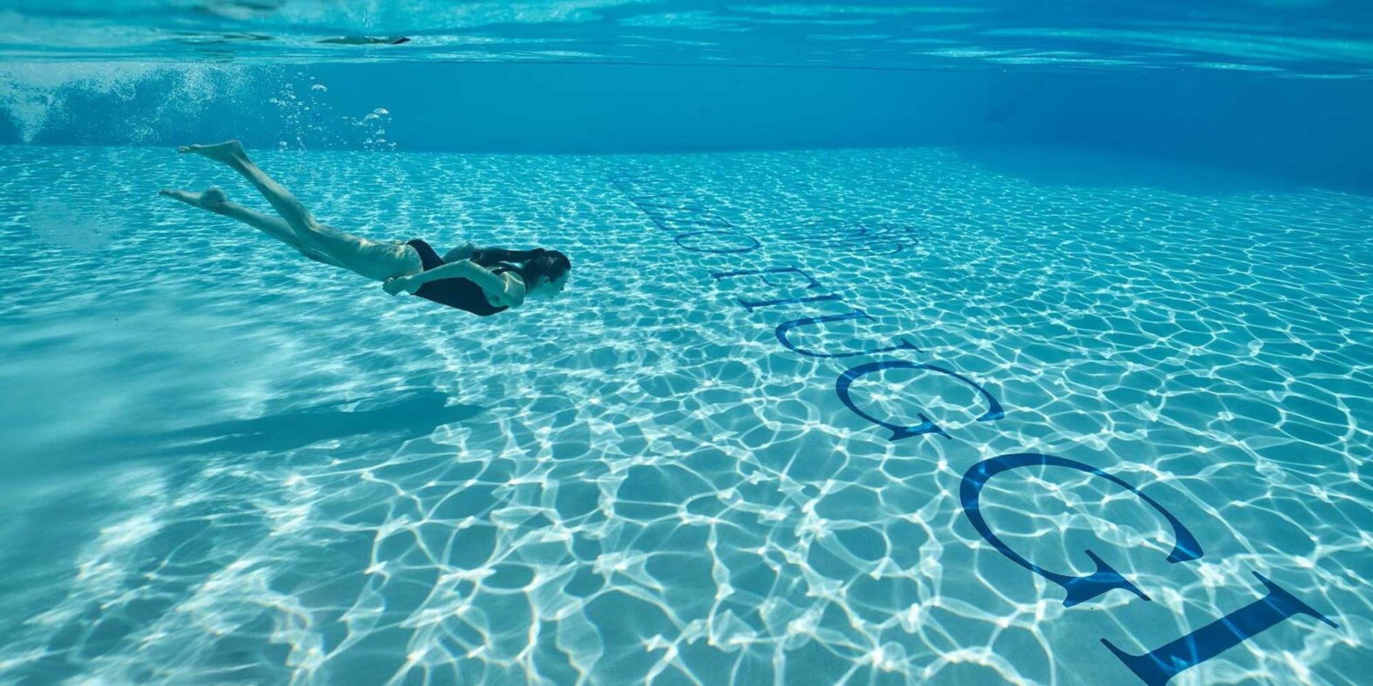 Person swimming underwater in a clear blue pool, side view, with sunlight patterns on the pool floor, bubbles ahead.