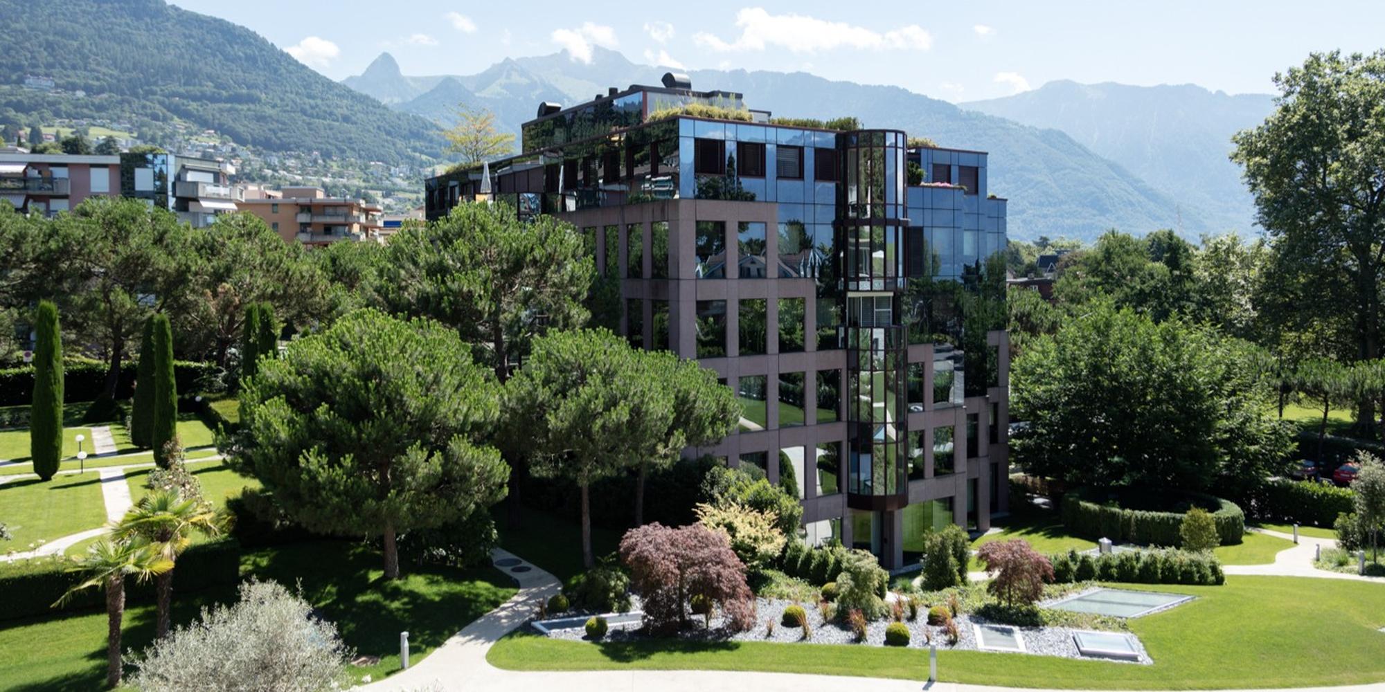 Modern glass-clad office building surrounded by manicured gardens and trees with mountains in the background.