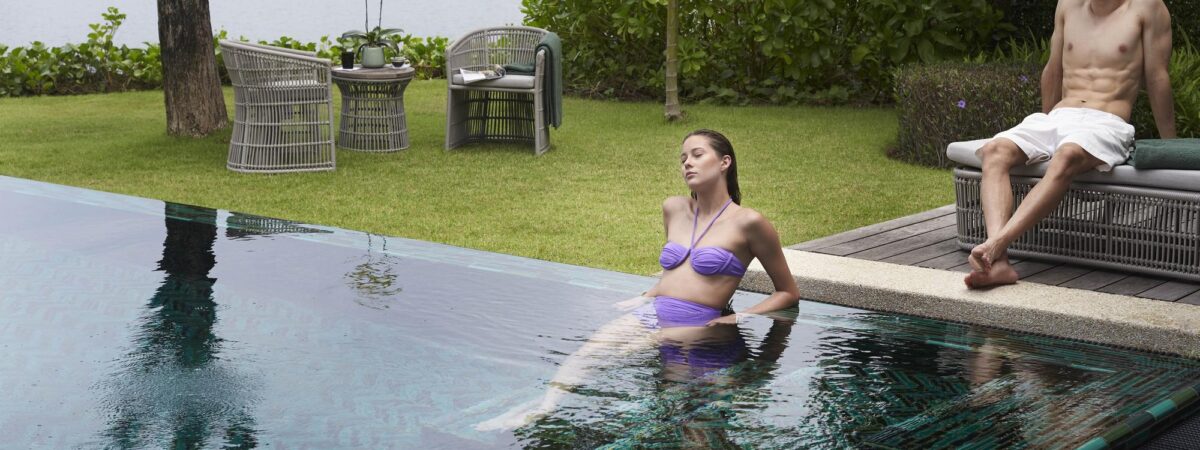 Woman in a purple bikini relaxing in a swimming pool near a man seated on a lounge chair by the poolside, with greenery and lake in the background.