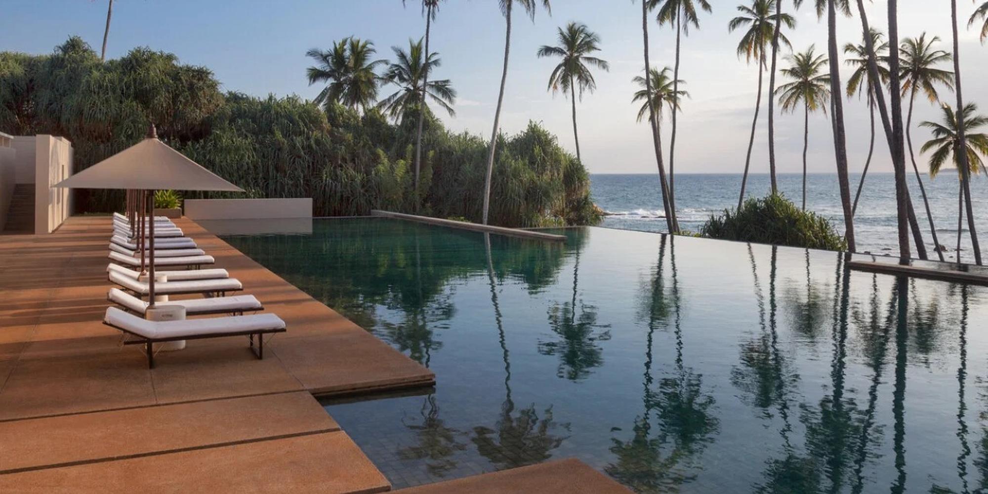 Infinity pool with a wooden deck, white lounge chairs under a large umbrella, and palm trees overlooking the ocean.