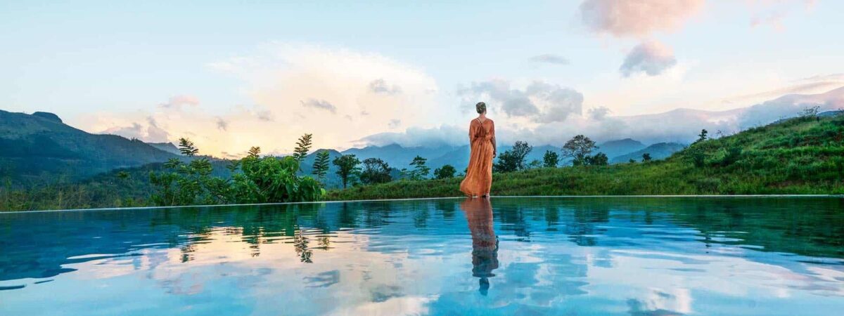 Woman in an orange dress standing at the edge of a reflective infinity pool, overlooking a mountainous landscape at sunset.