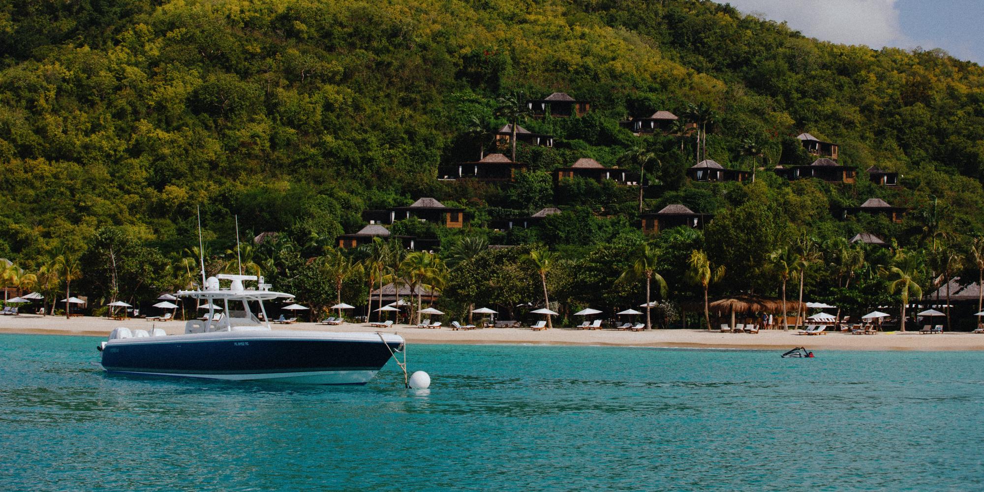 Large motorboat anchored near a sandy tropical beach with palm trees, beach umbrellas, and hillside huts in a green forested backdrop.