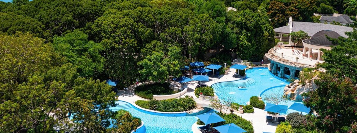 Resort pool area surrounded by palm trees and blue umbrellas, ocean visible in the distance.
