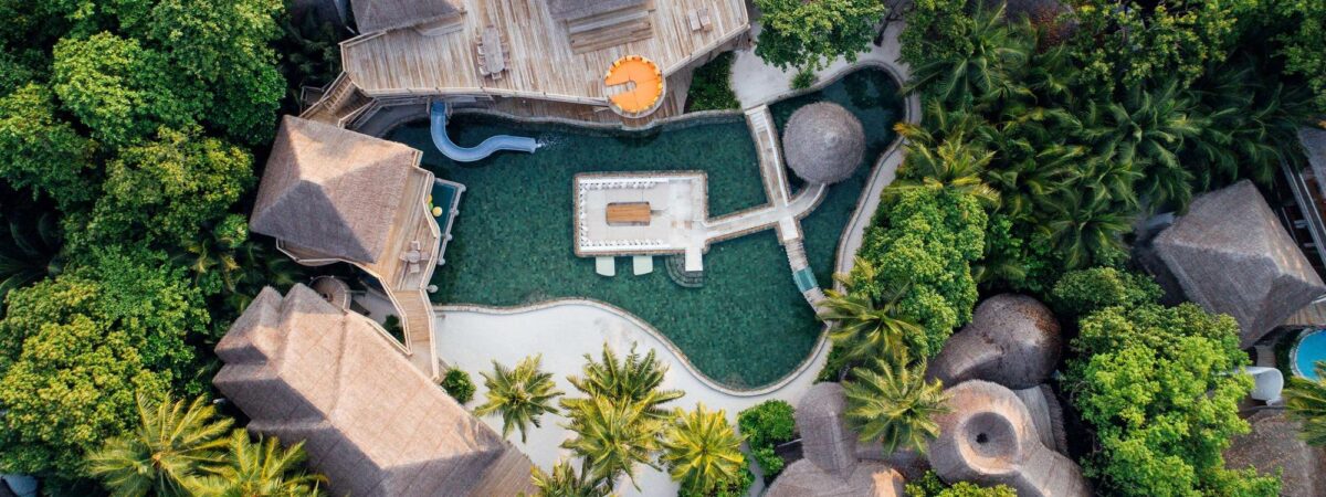 Aerial view of a tropical resort with thatched-roof buildings, a winding pool, and lush green trees around.