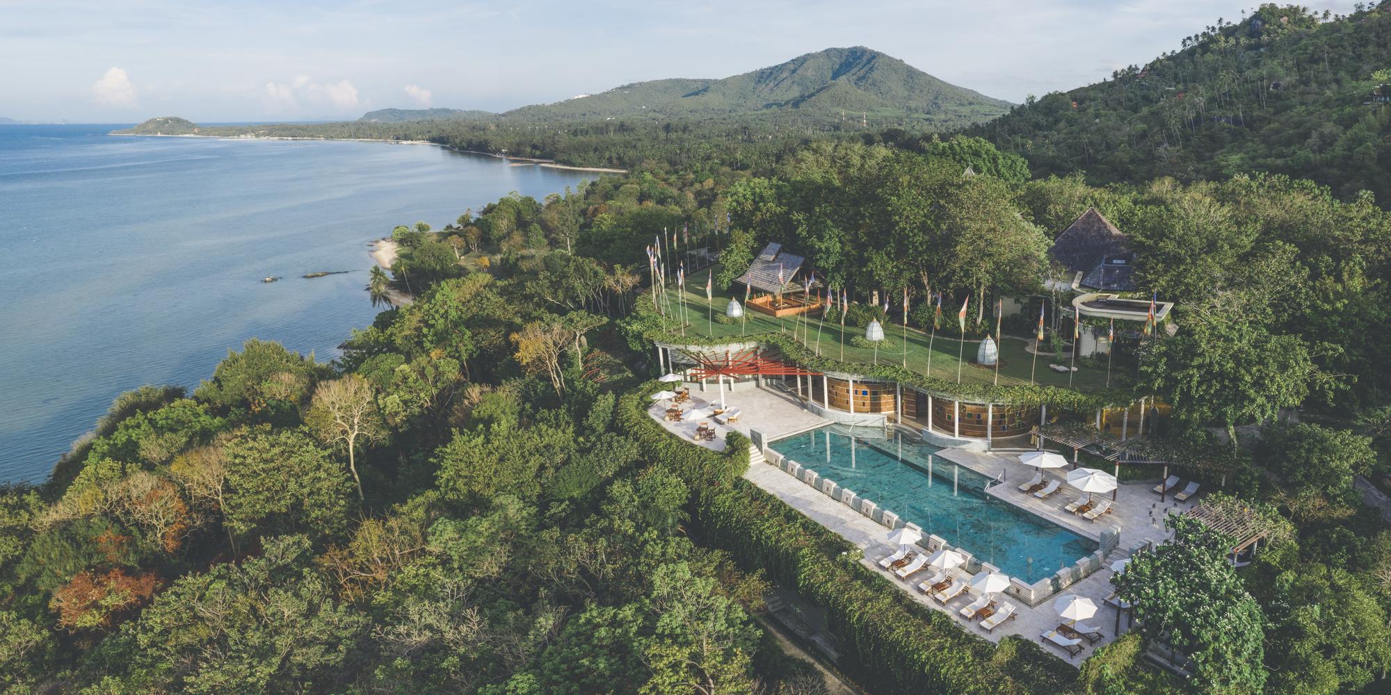 A tropical resort with a long pool, sun loungers and umbrellas among lush greenery overlooking the sea.