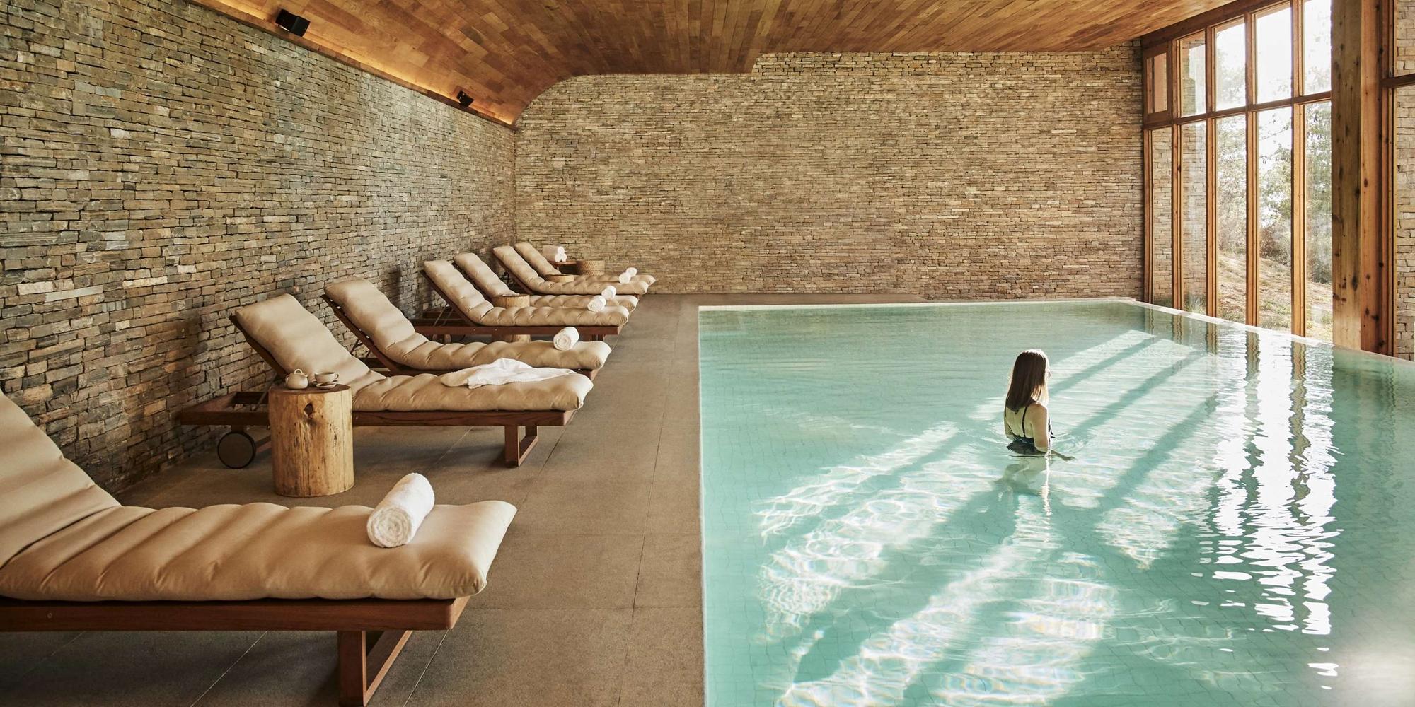 Indoor spa pool with stone walls, beige lounge chairs along the edge, and towels, by large wooden-framed windows.