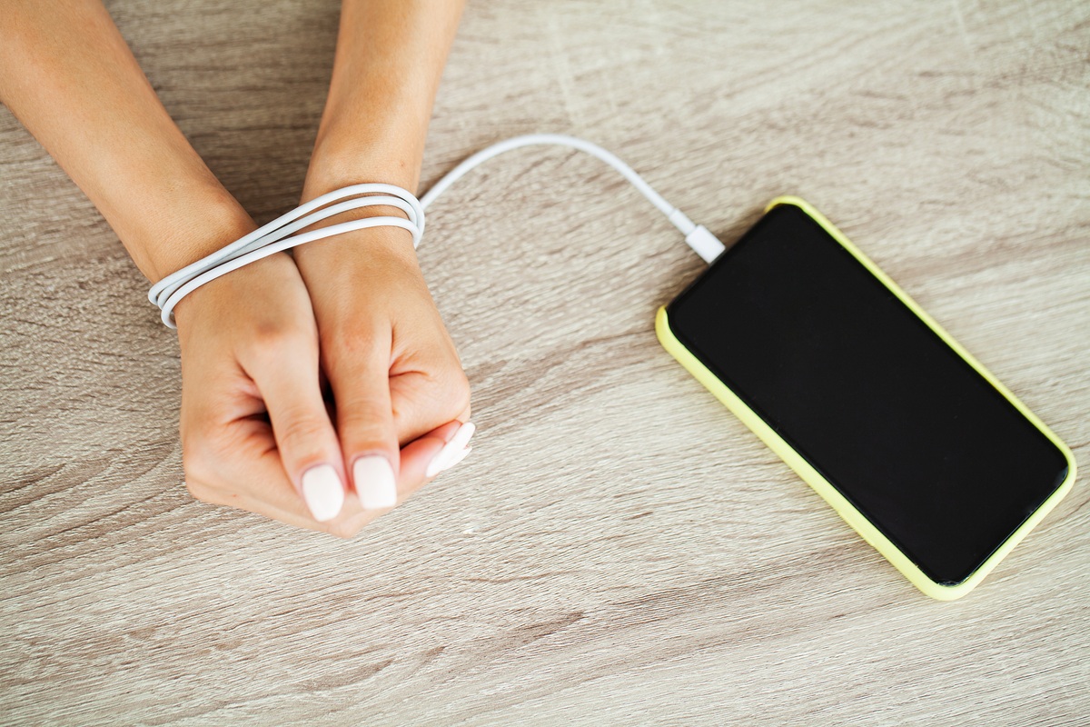 Hands tied with a phone charging cable next to a smartphone on a wooden surface, symbolizing technology or phone addiction.39% of adults are overwhelmed, according to the American Psychological Association, with perpetual messages and notifications. Hands tied with a phone charging cable next to a smartphone on a wooden surface, symbolising technology- or phone addiction.