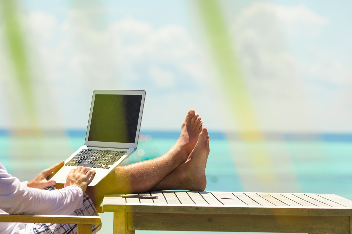 Person relaxes on a wooden deck by the turquoise ocean, using a laptop. Sunlight filters through palm leaves, creating a peaceful, tropical setting. These digital detox retreats are among the best in the world.