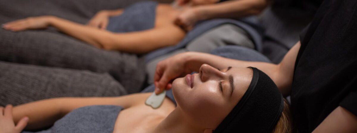 Two clients lie on massage beds with eyes covered by towels or eye pillows, receiving spa treatments in a dim room