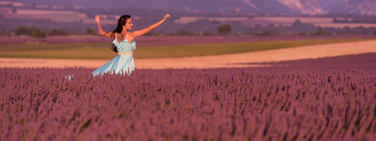 Woman in a light blue dress running through a lavender field with distant hills behind her.]?