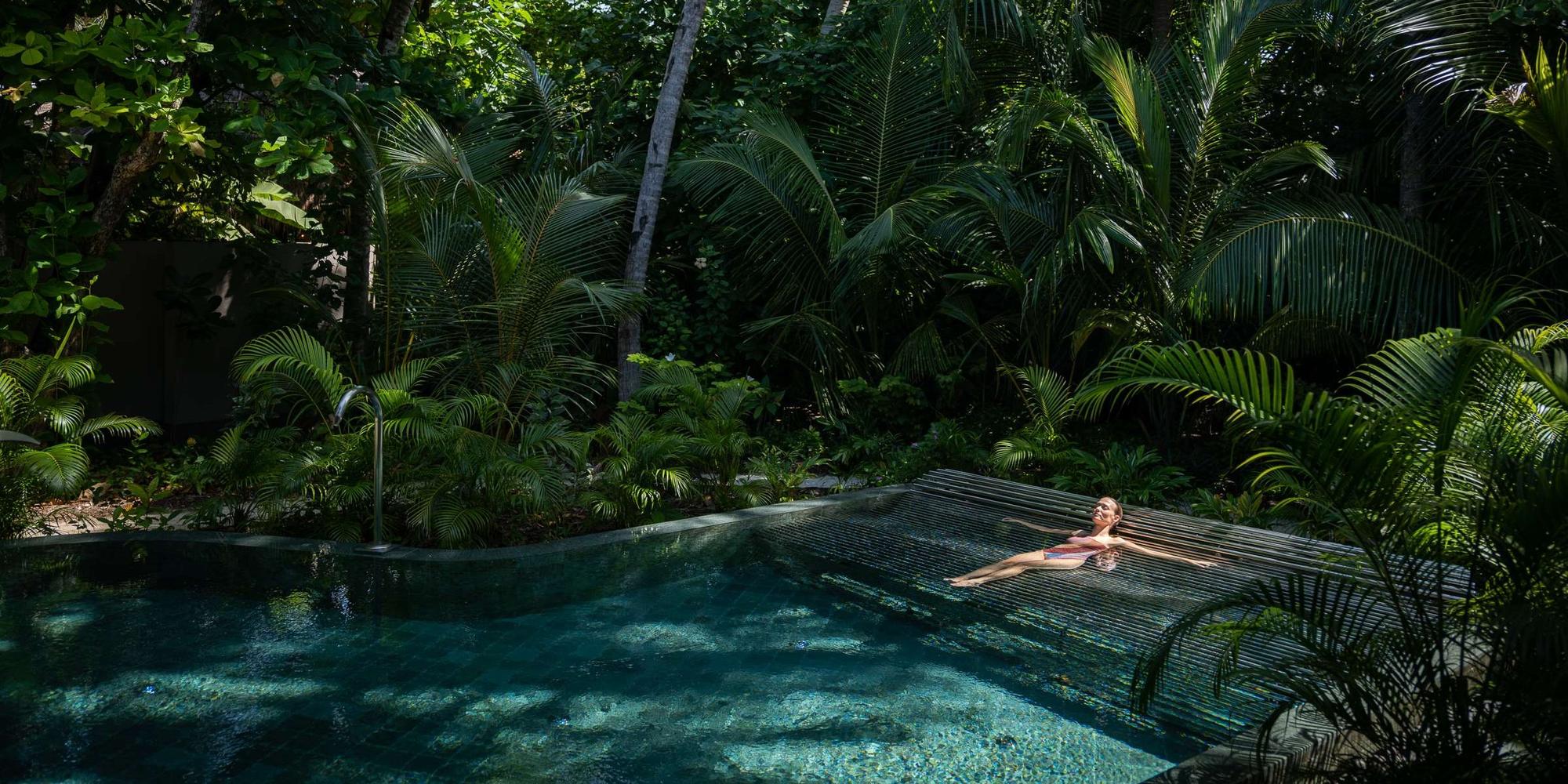 A tranquil tropical pool area featuring a sparkling blue pool with a woman inside the pool at Joali Being. A tranquil tropical pool area featuring a sparkling blue pool with a woman inside the pool at Joali Being.