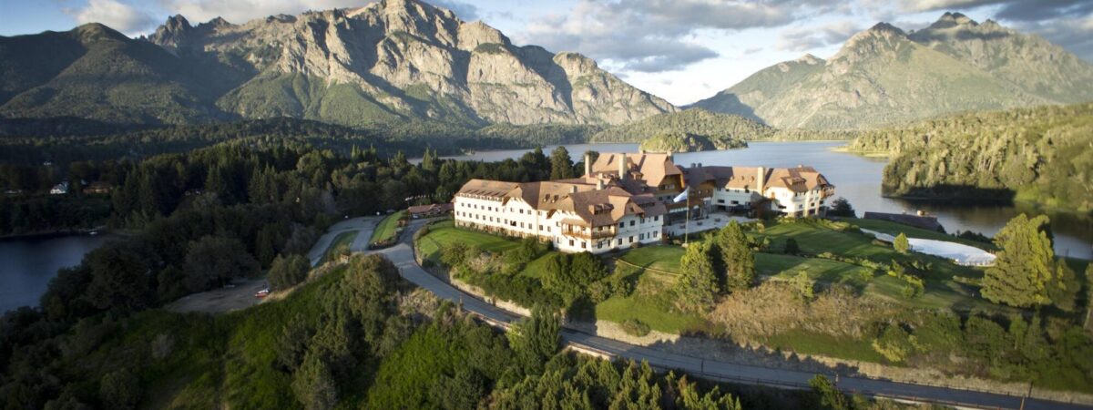 Alpine lodge complex beside a lake with forest and rugged mountains in the background, viewed from above.