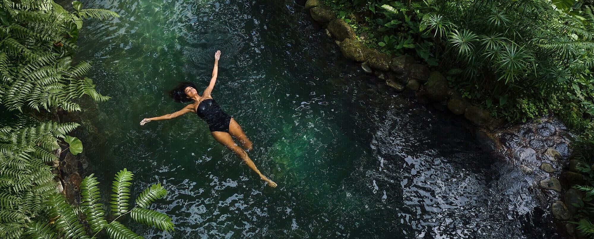 A woman floating in a dark natural body of water surrounded by lush foliage and ferns. A woman floating in a dark natural body of water surrounded by lush foliage and ferns.
