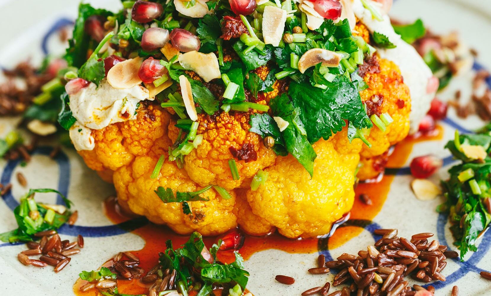 A vibrant whole roasted cauliflower dish, likely a spiced or curry-roasted head, garnished with fresh herbs, pomegranate seeds and a creamy sauce or yogurt base. A vibrant whole roasted cauliflower dish, likely a spiced or curry-roasted head, garnished with fresh herbs, pomegranate seeds and a creamy sauce or yogurt base.