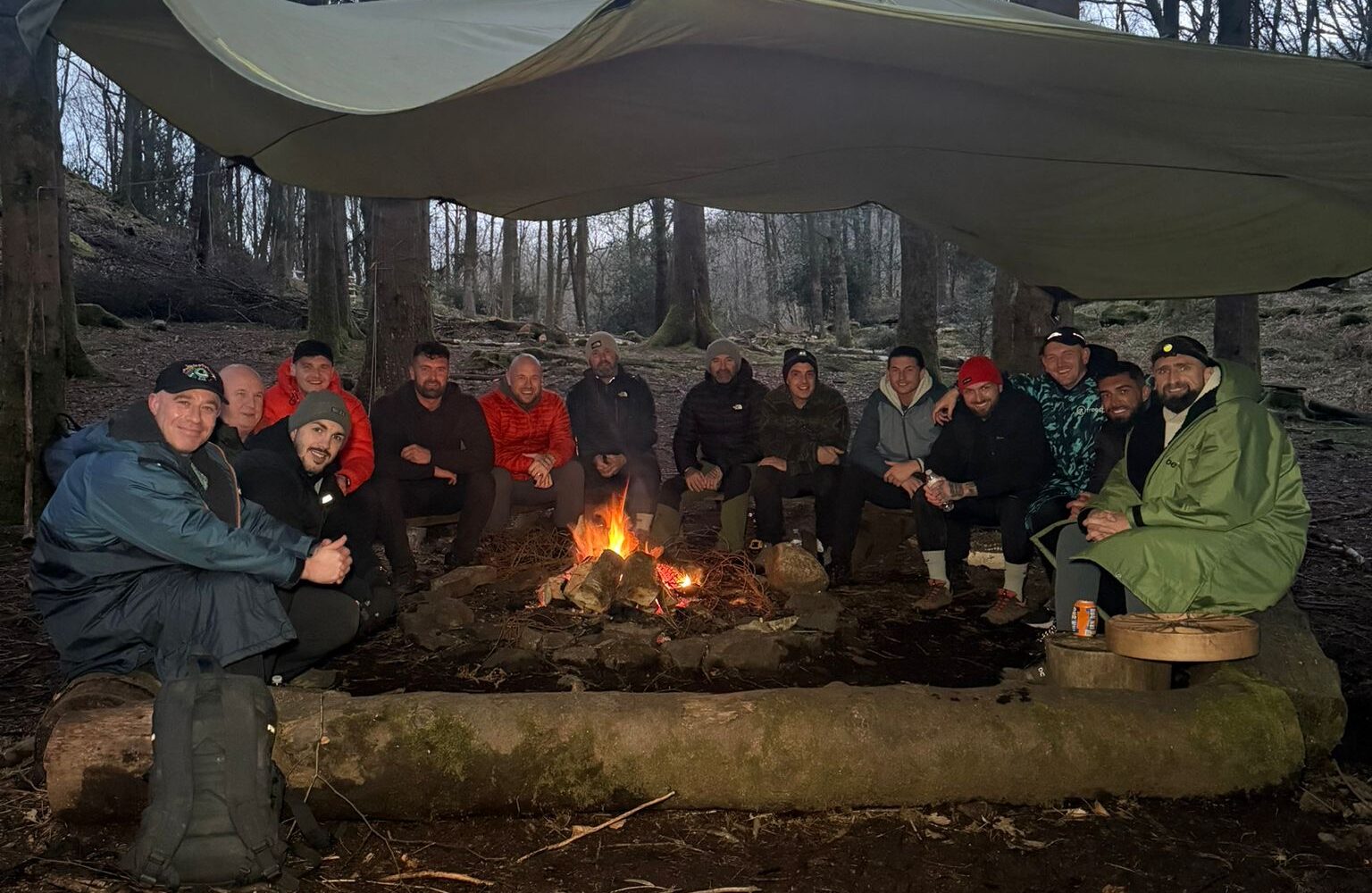 The image showing a group of men gathered around a campfire in a wooded area beneath a tarp or shelter.