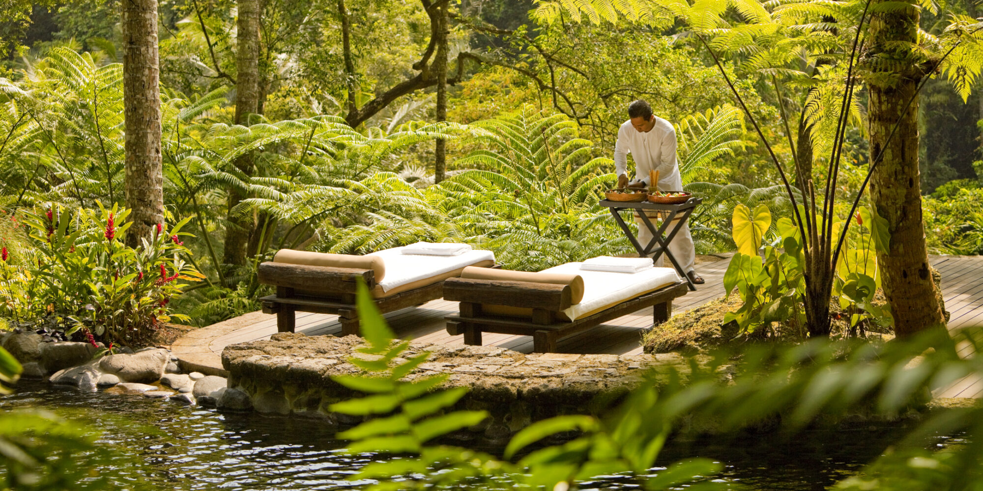 A tranquil outdoor wellness setup with wooden massage loungers beside a water feature. A tranquil outdoor wellness setup with wooden massage loungers beside a water feature.